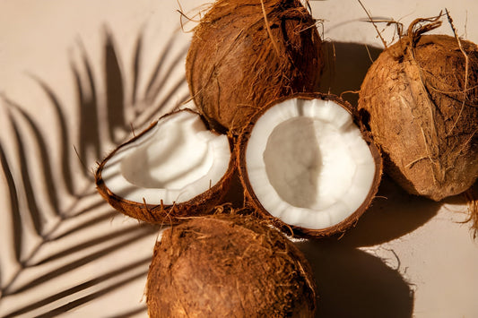 Fresh coconuts, some cut open to show the white meat, with a tropical palm leaf shadow on a light background, symbolizing natural coconut oil